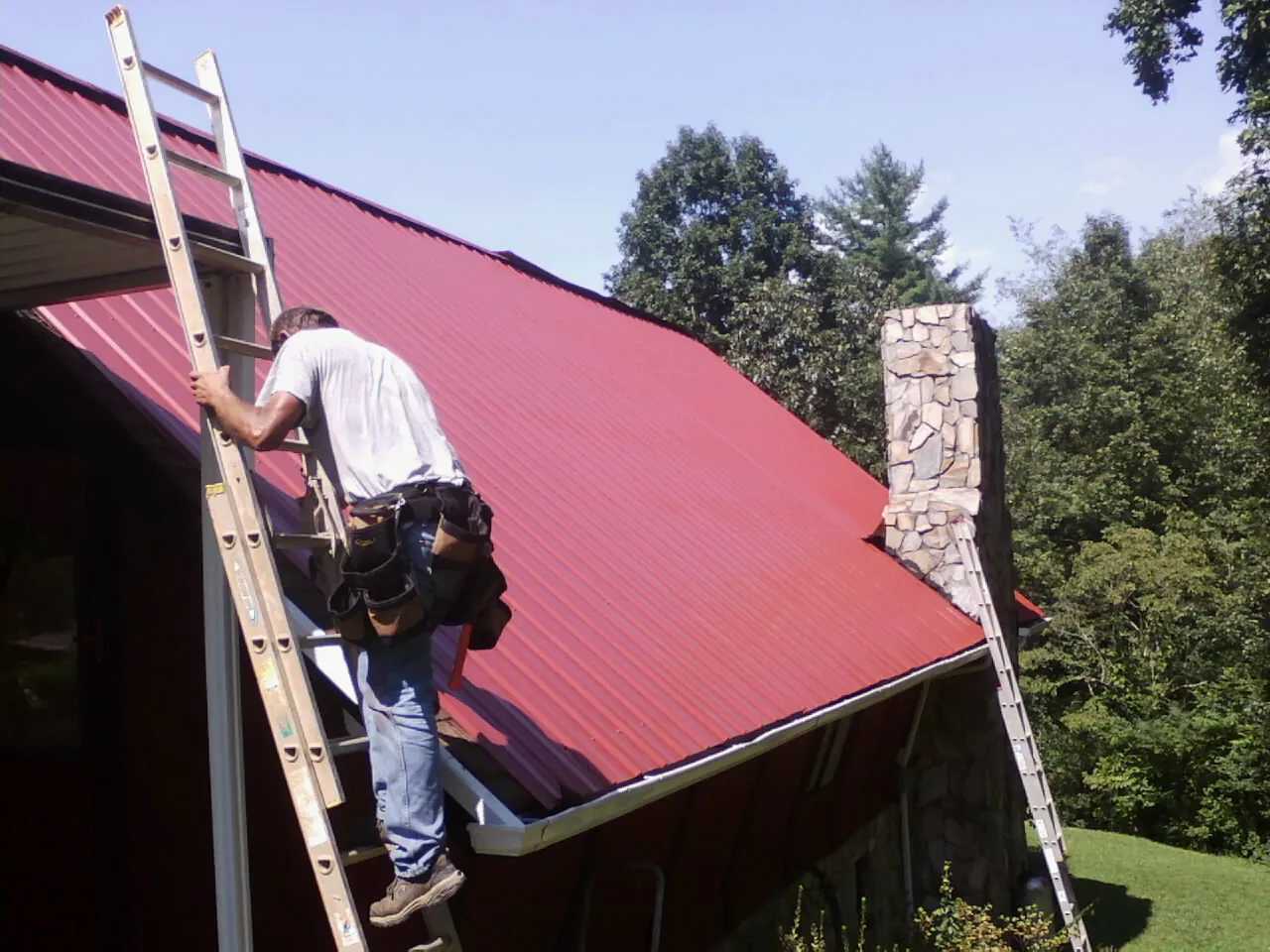 Roofer on a ladder beside a red metal roof and stone chimney — Soffit & Fascia Repair by Roofing Brothers in Winterset